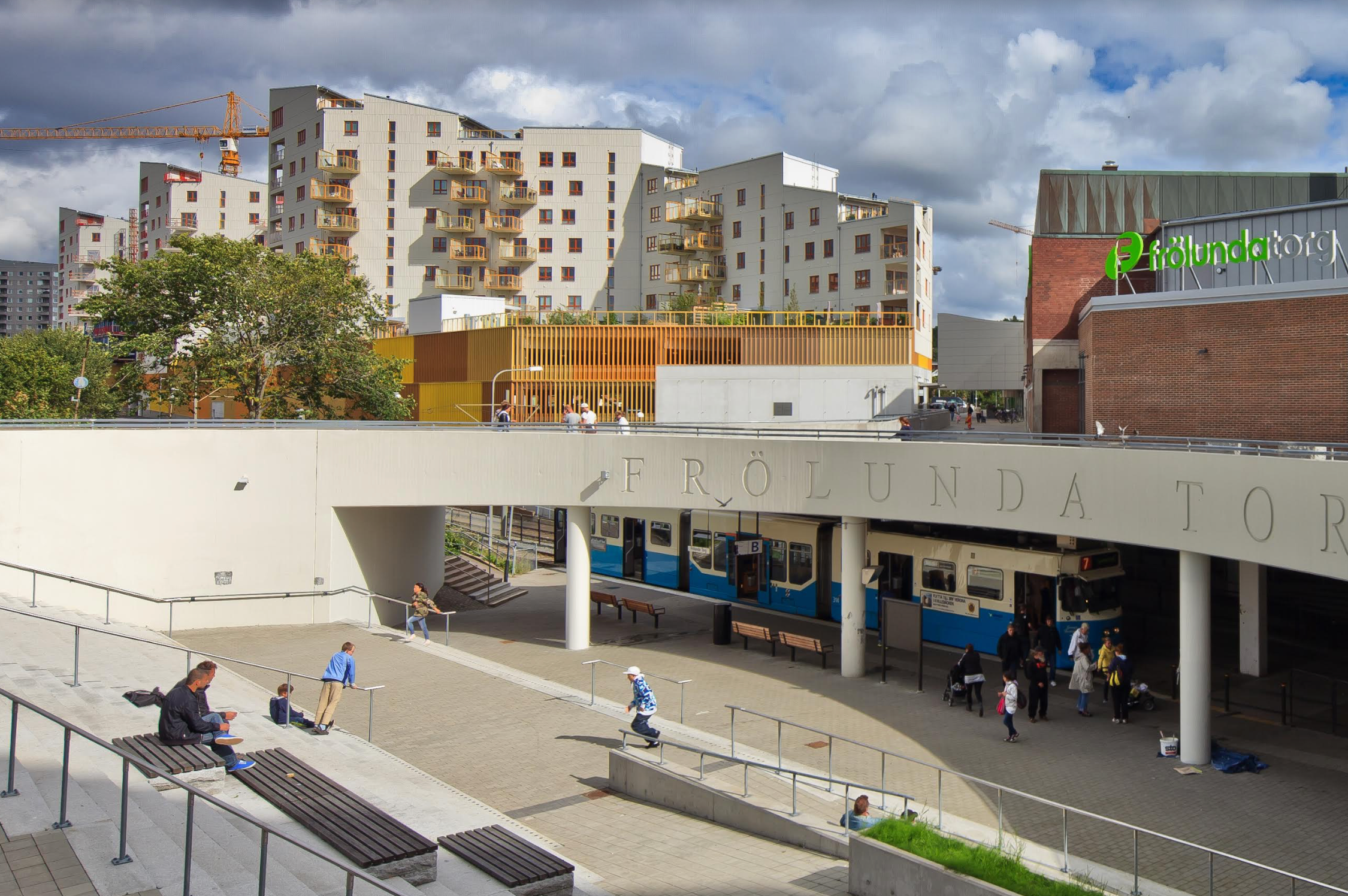 Foto på Frölunda Torg i Göteborg. Människor sitter i trappen, spårvagnen passerar.
